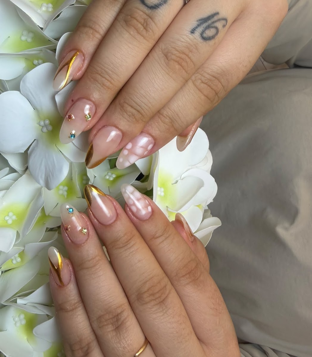 Dos manos con uñas almendradas en cromo dorado, con flores y piedras, sobre orquídeas blancas