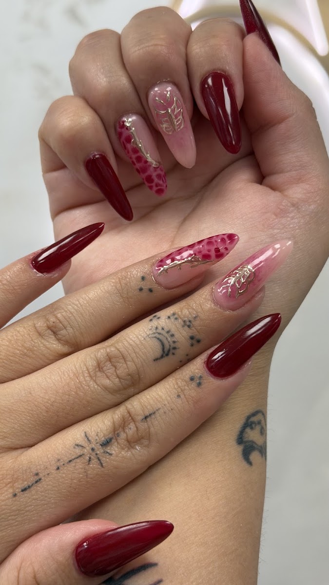 Manos con uñas stiletto largas en burdeos y nude, con flores pintadas y detalles dorados; se ven tatuajes en los dedos