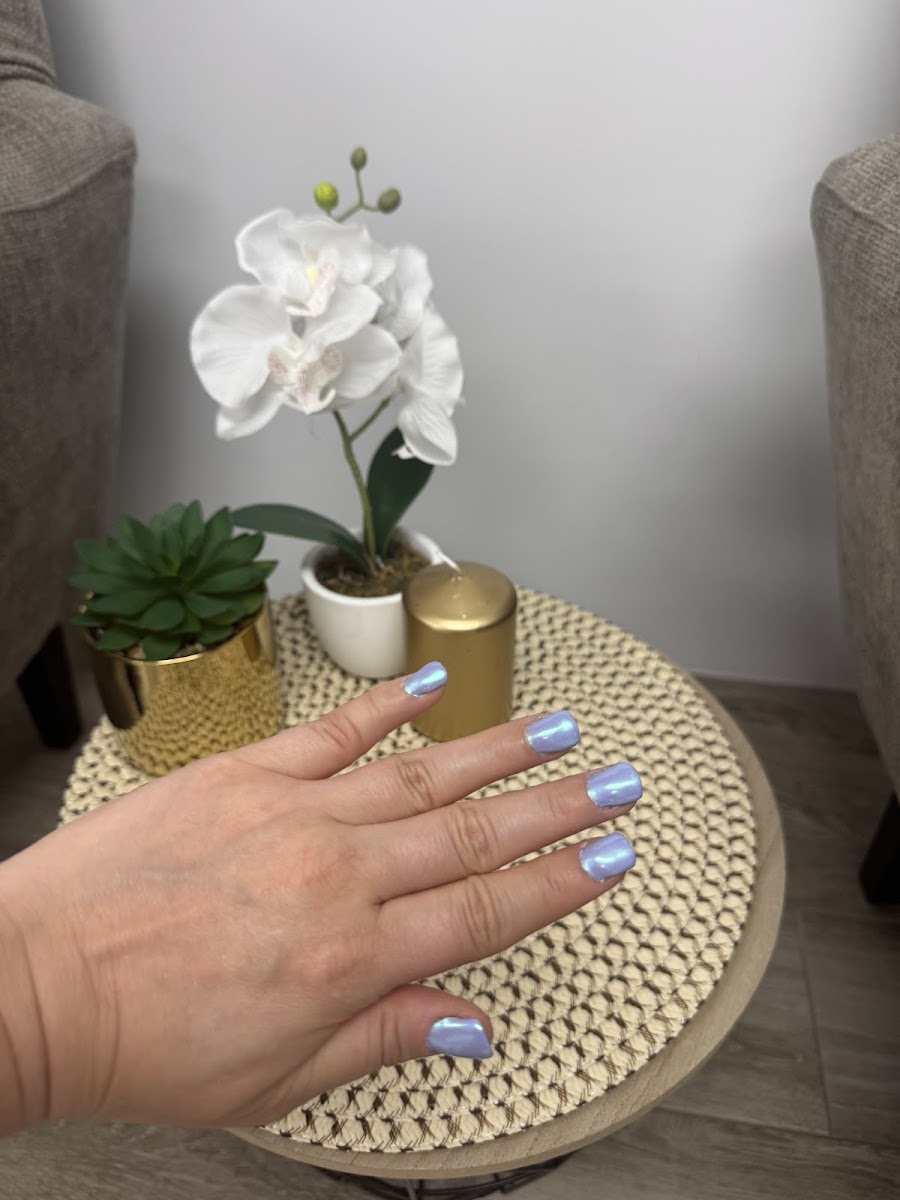 Mano con esmalte violeta iridiscente sobre un mantel de mimbre, junto a una orquídea blanca y una maceta dorada