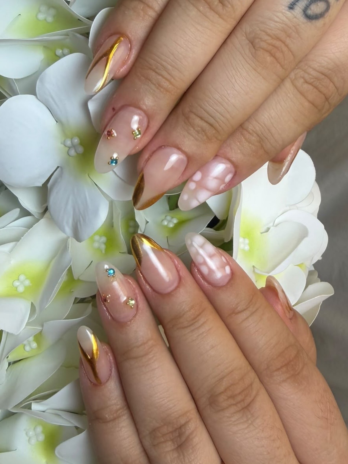 Dos manos con uñas almendradas decoradas con dorado y pedrería sobre flores blancas